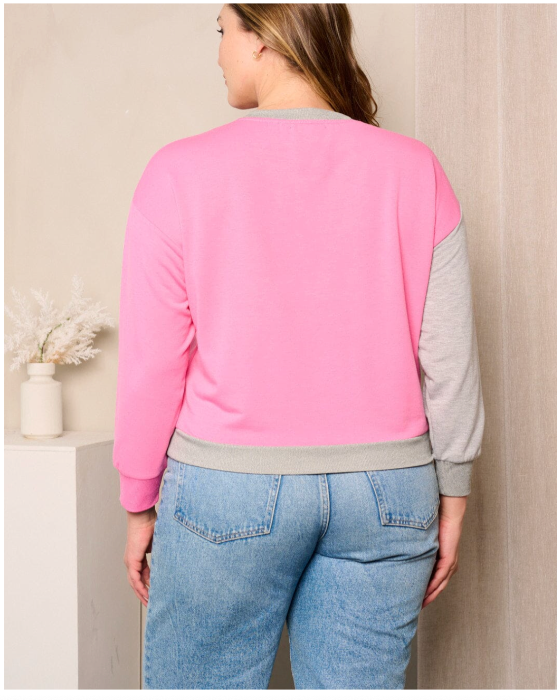 Person wearing a pink and gray sweater with blue jeans against a neutral wall.