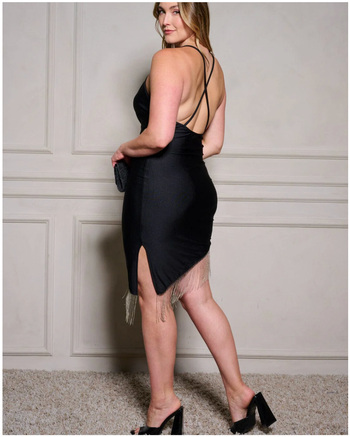 Woman wearing a black dress with thin straps and a slit, standing against a neutral wall.